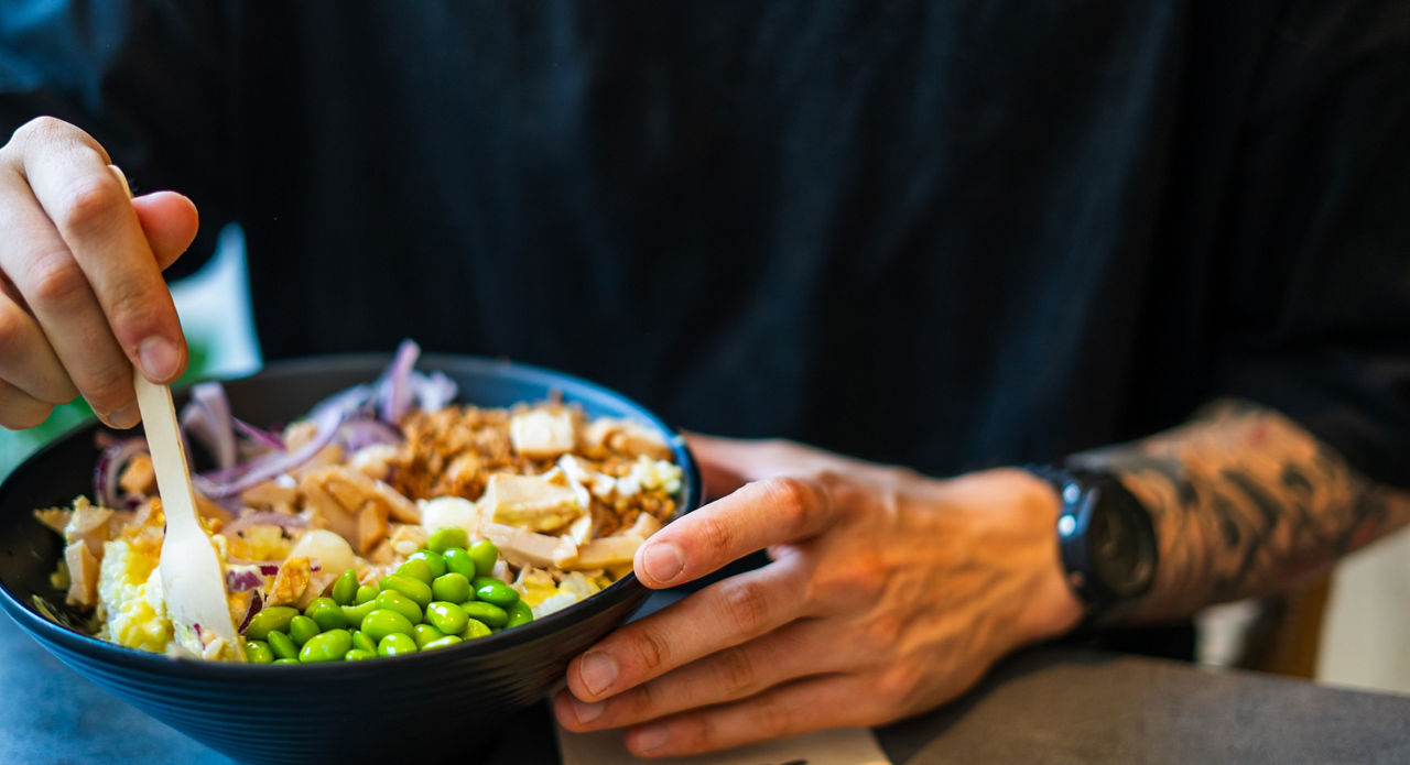 young tattooed man holding a poke bowl on a black plate and eating it